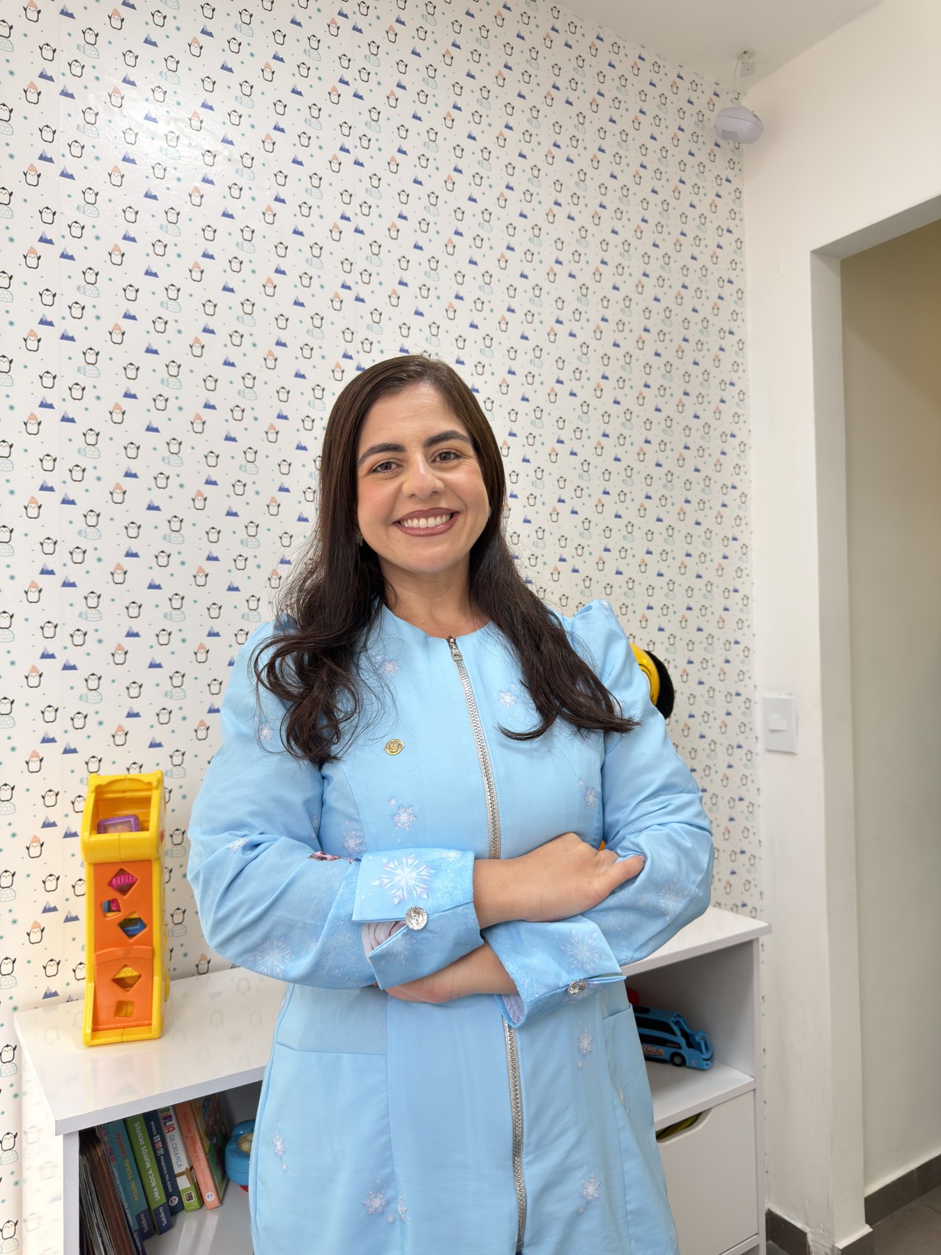 Dra. Iony sorrindo no espaço kids da clínica OdontoDreams, com papel de parede de pinguins e brinquedos.